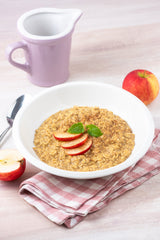 Bowl of oatmeal with sliced apples on a checkered cloth, next to a purple pitcher and apple slices.